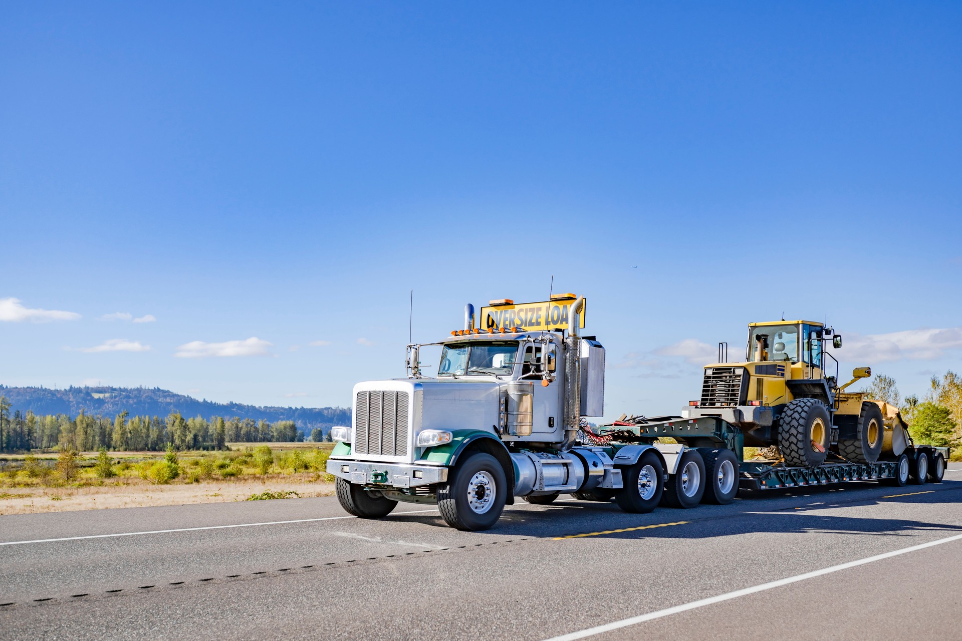 Powerful white day cab big rig semi truck with oversize load sign on the roof transporting heavy tractor on step deck semi trailer running on the highway road